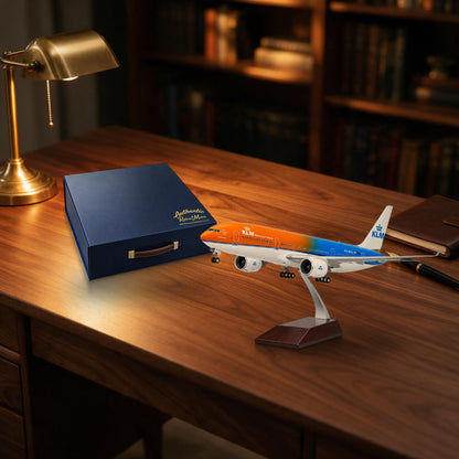 A miniature KLM Royal Dutch Airlines model airplane on a display stand, placed on a wooden desk next to a dark blue presentation box and a brass desk lamp, with blurred bookshelves in the background.
